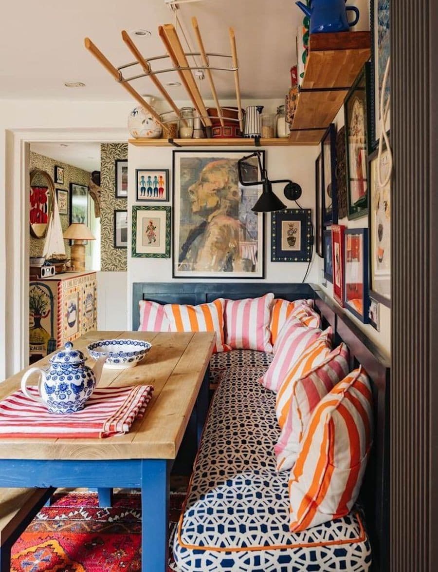 Comedor de estilo cluttercore con una banca con cojines de rayas rosadas y anaranjadas, una mesa de madera con patas pintadas azules, paredes llenas de cuadros de distintos tamaños. Sobre la mesa hay una tetera y plato pintados con un diseño azul.