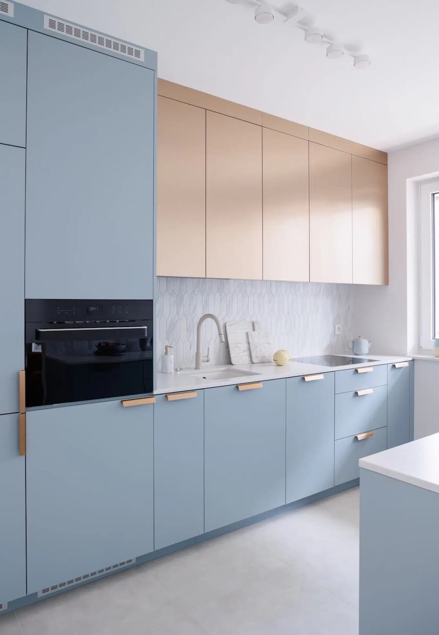 Cocina con muebles de color azul bruma, cubierta y pisos blancos, uno de los muebles superiores es de madera clara.