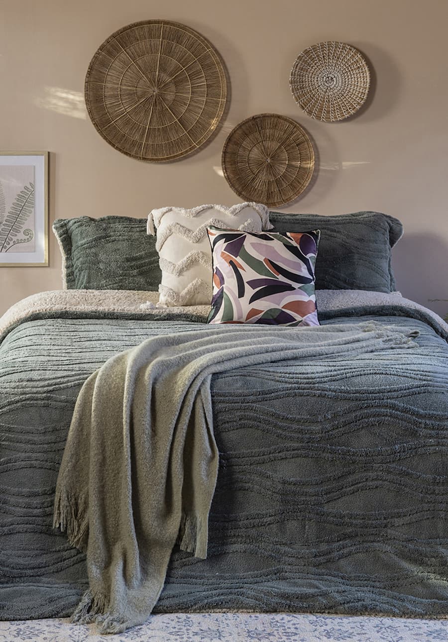 Dormitorio de estilo Boho chic con una cama con un plumón verde oscuro, una manta verde pastel, cojines verdes, blancos y uno de colores tierra. En la pared del fondo hay 3 platos de fibras naturales.