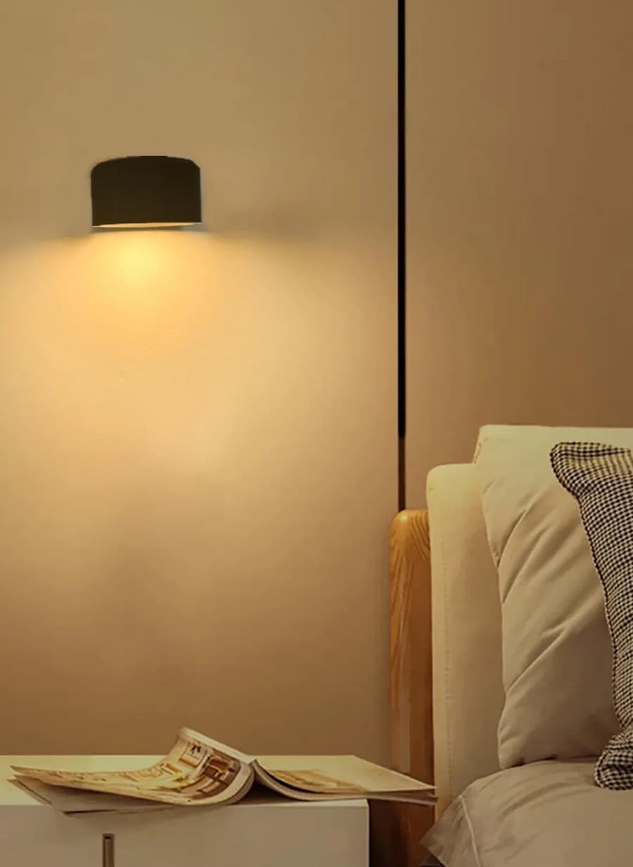 Detalle de un dormitorio con una cama de madera y sábanas blancas, un velador y sobre él, un apliqué de muro que entrega una luz cálida y relajante al espacio. 