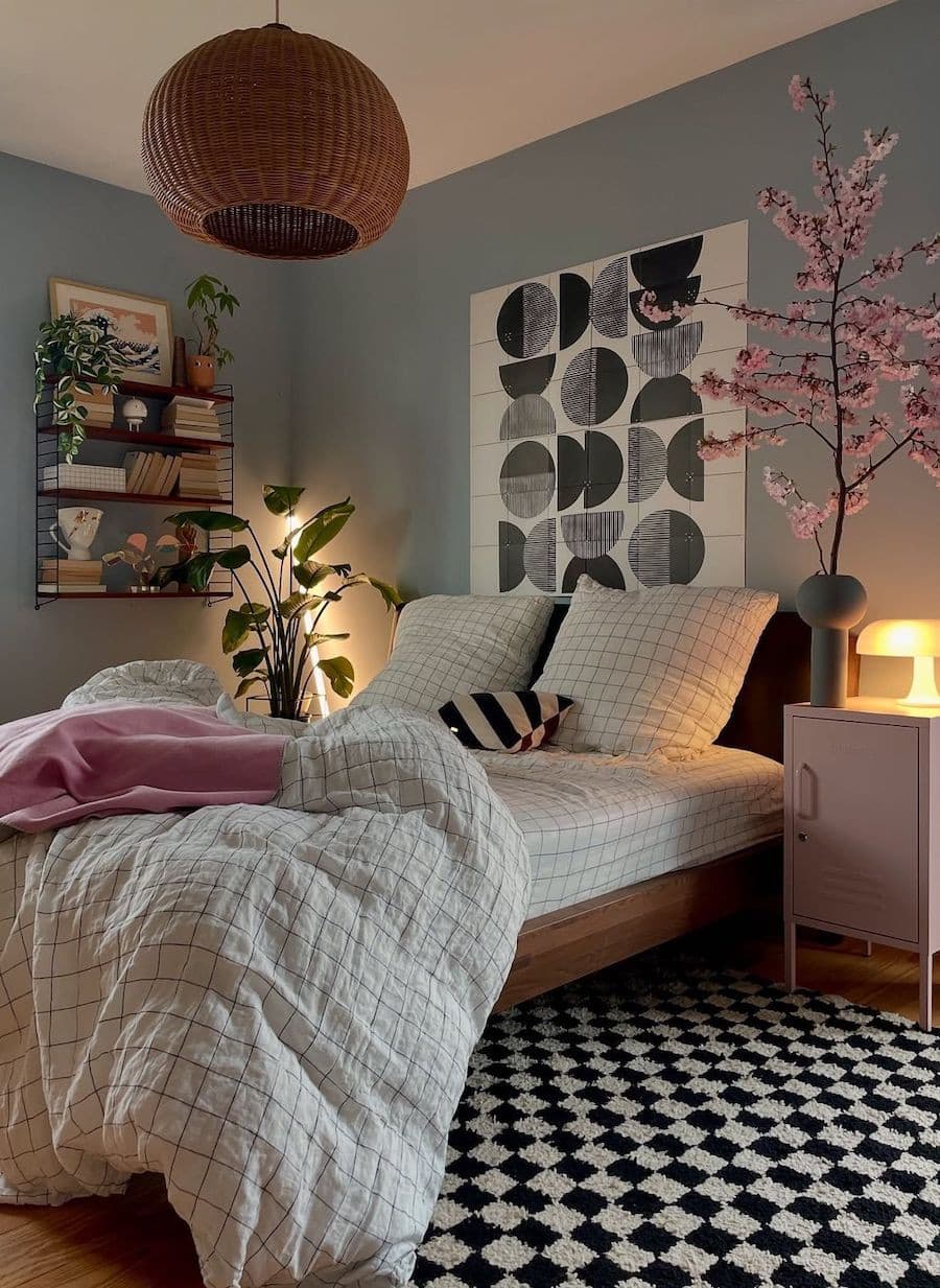 Dormitorio de estilo cozy con paredes celeste grisáceo, cama de madera, ropa de cama blanca con cuadros de líneas negras, una manta y velador rosados, iluminación tenue desde 2 lámparas, una alfombra de cuadros en blanco y negro.