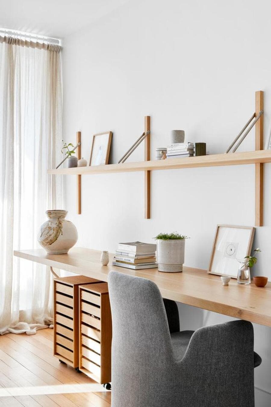 Escritorio minimalista. Mesa de madera flotante con dos cajoneras bajo ella. Silla de escritorio gris. Repisa flotante de madera sobre un muro blanco. Jarrones de cerámica, libros y adornos. Gran ventana con cortina de tela vaporosa y blanca