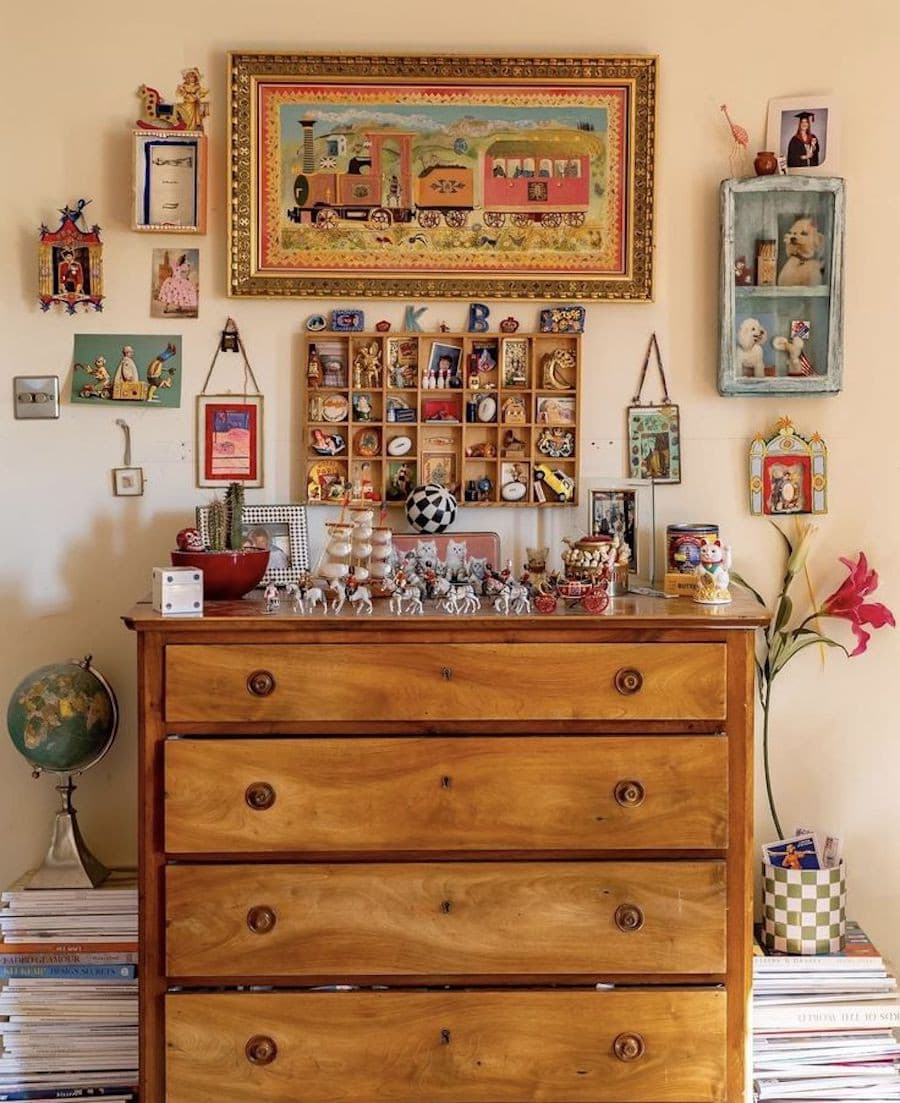 Pared color crema con cuadros de diferentes estilos, adelante hay una cómoda de madera y sobre ella muchos adornos y recuerdos de viajes de muchos colores.