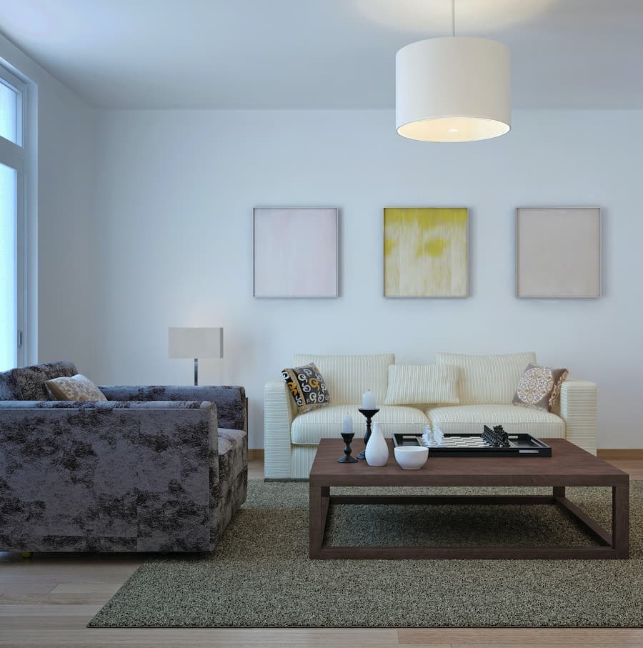 Living de paredes blancas, una gran lámpara colgante con pantalla redonda y blanca, un gran sofá blanco y un sofá de un cuerpo con estampado gris oscuro, una mesa de centro oscura, una alfombra verde musgo, en la pared hay 3 cuadros de colores pastel.