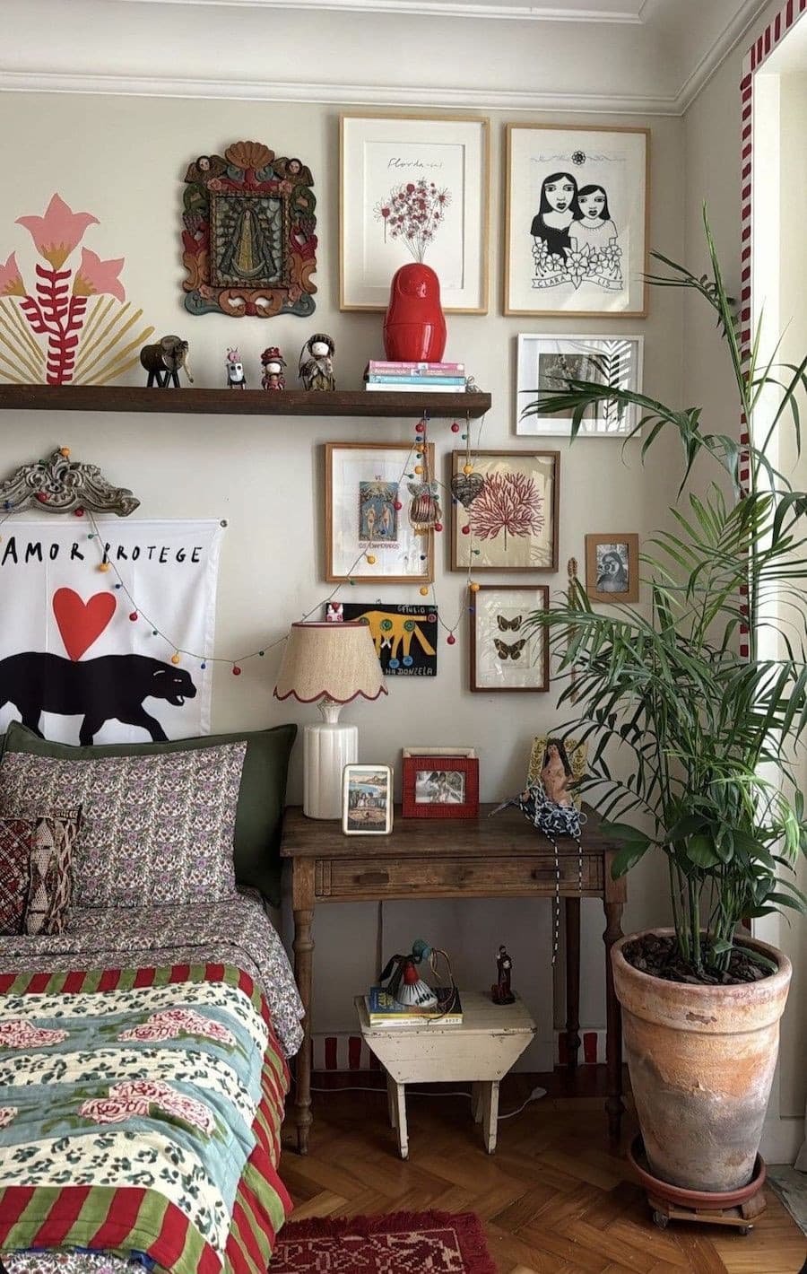 Dormitorio de estilo cluttercore con un cama con cobertor estampado de colores oscuros, un velador de madera, pequeños cuadros en las paredes, una gran planta.