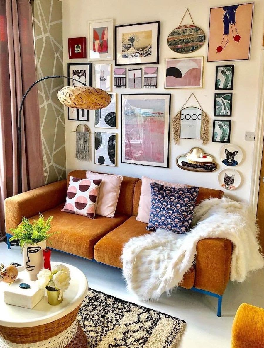 Living con una sofá de color camel con cojines con estampados rosados sobre él y una manta peluda blanca. En la pared del fondo hay un muro galería con distintos cuadros con ilustraciones y arte colorido y con significado.