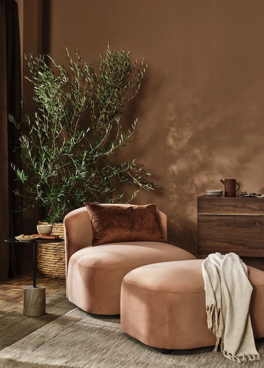Detalle de una sala de estar con el color Pantone 2025, Mocha Mousse. Poltrona redonda y baja con apoyapiés de color café. Atrás hay un olivo en un canasto. Muros de color café