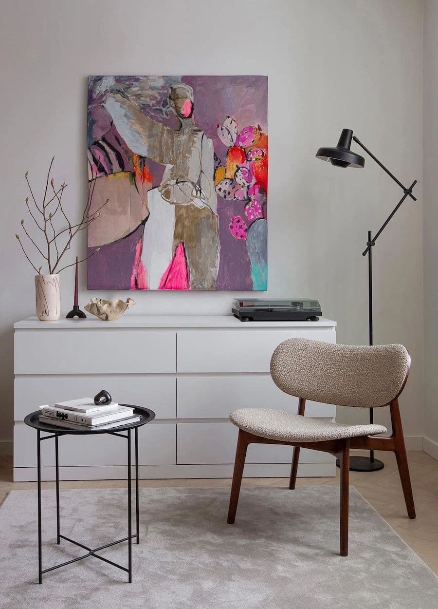 Sala de estar con una cómoda blanca tipo buffet, una lámpara de pie negra de forma irregular, una silla de madera oscura con tapiz beige, una mesa lateral negra con libros. Y al fondo, un gran cuadro protagonista de colores flúor y formas abstractas.