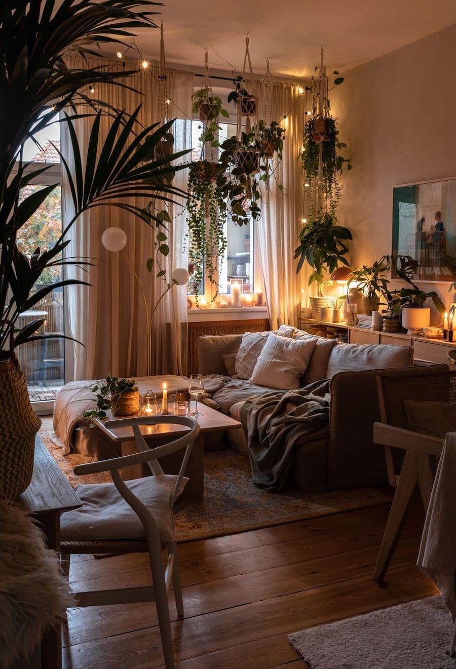 Sala de estar de estilo cozy con varias fuentes de iluminación tenue, un sofá beige con mantas verde oliva, una alfombra mullida y muchas plantas alrededor.