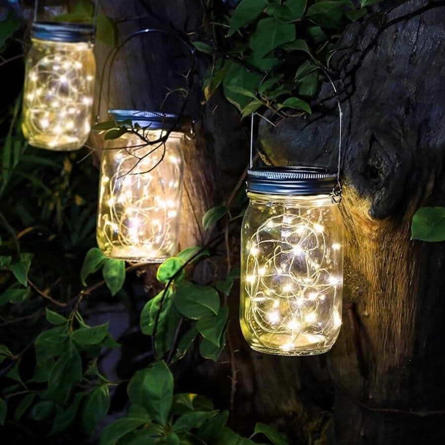 Tres frascos de vidrio, con tapa metálica y con guirnaldas de luces en su interior. Están colgados en un muro de madera, con plantas enredaderas a su alrededor.