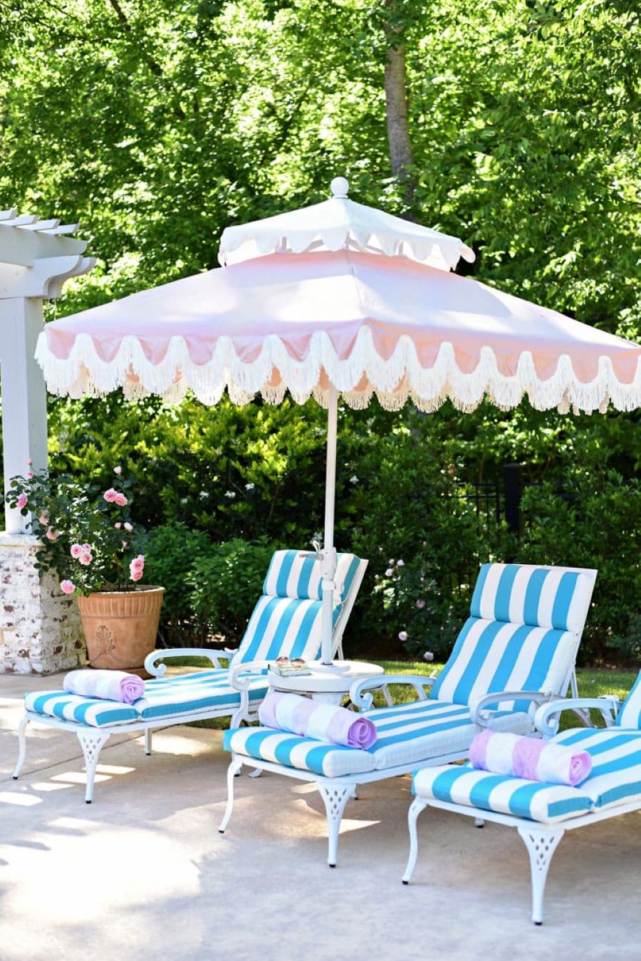 Tres reposeras con cojines con rayas celestes y blancas. Gran quitasol cuadrado, de estilo vintage, rosado y blanco. Sobre las reposeras hay toallas enrolladas. Atrás hay un macetero de arcilla con una planta y muchos árboles.