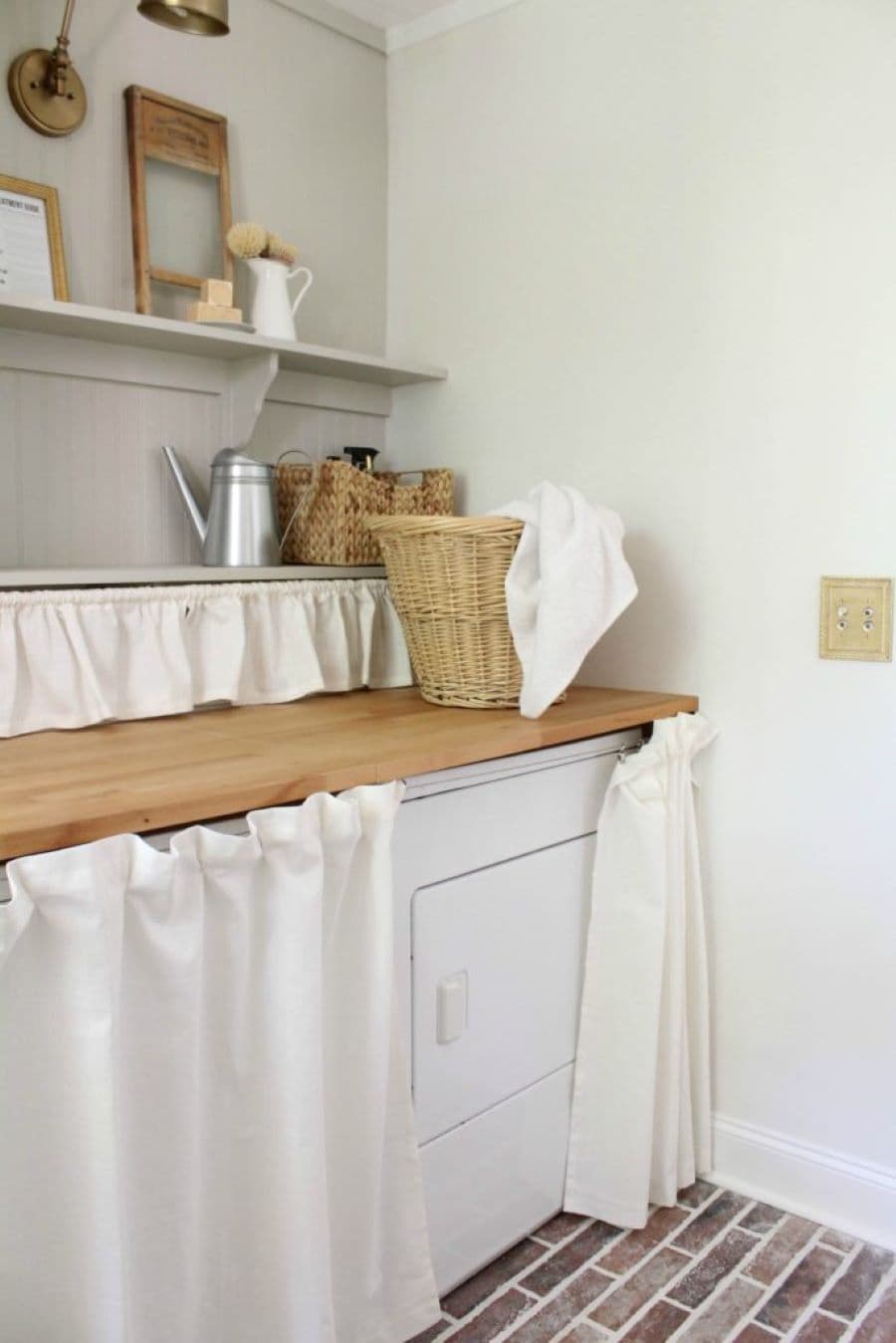 Logia con lavadora escondida detrás de cortina color blanco. Mesón de madera clara, con muro con revestimiento de madera gris. Repisa flotante con una caja de mimbre y artículos de aseo. Canasto de mimbre alto con una tela blanca