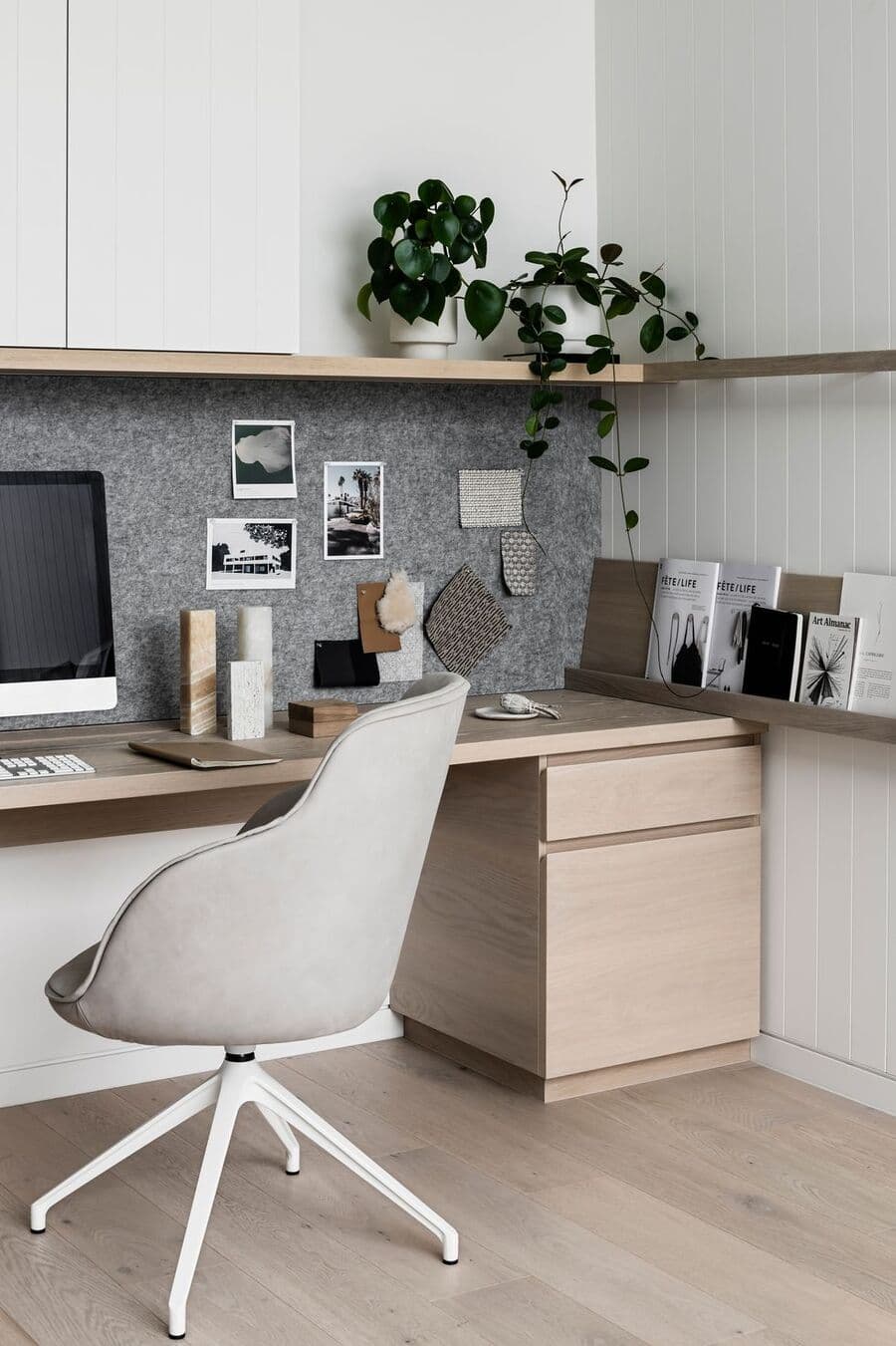 Escritorio clásico con mueble de madera pegado al muro. Una cajonera de madera, silla de escritorio con ruedas, color gris claro. Pared gris con dos repisas flotantes de madera con revistas y plantas. Muro blanco con revestimiento de madera.