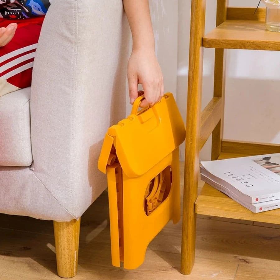 Mano sosteniendo un piso de plástico plegable amarillo. Se ve la esquina de un sillón color beige con patas de madera y una repisa de madera con unas revistas en el nivel inferior. Piso de madera.